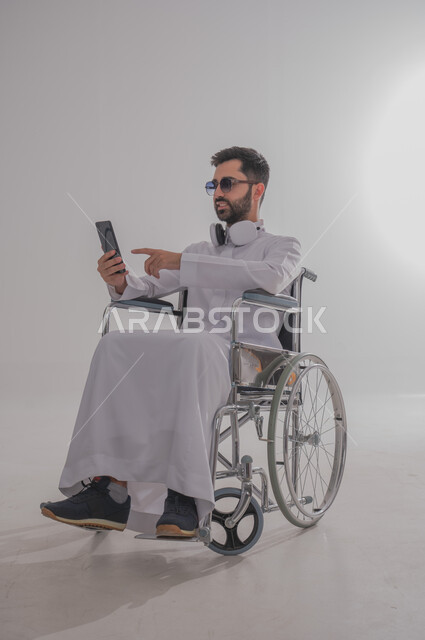 Holding a mobile phone, browsing the internet and knowing the latest news and developments, portrait of a young Saudi Arabian Gulf man with special needs wearing traditional dress and sunglasses sitting in a wheelchair and wearing headphones, using modern techniques and technology, gray background