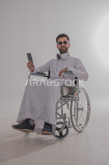 الإمساك بالهاتف المحمول وتصفح مواقع الانترنت ومعرفة آخر الأخبار والمستجدات، بورتريه لشاب عربي خليجي سعودي من ذوي الاحتياجات الخاصة يرتدي الثوب التقليدي والنظارات الشمسية يجلس على كرسي متحرك ويضع سماعات الأذن، استخدام التقنيات والتكنولوجيا الحديثة، خلفية رمادية