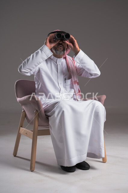 مفهوم رؤية الأشياء البعيدة، بورتريه لرجل عربي خليجي سعودي يرتدي الثوب التقليدي والشماغ يجلس على كرسي مريح  ينظر من خلال التلسكوب, حلم تحقيق الأهداف والطموحات المستقبلية, الشغف وحب الاستكشاف والمغامرة، البحث عن شيء ما باستخدام الناظور، خلفية رمادية