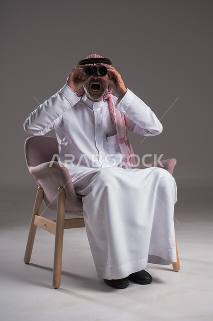 التخطيط المستقبلي للأهداف والطموحات، بورتريه لرجل عربي خليجي سعودي يرتدي الثوب التقليدي والشماغ يجلس على كرسي مريح  ينظر من خلال منظار الرؤية ثنائي العينين, حب الاستكشاف والمغامرة، رؤية الاشياء البعيدة عبر التلسكوب بايماءات الاندهاش والسرور، خلفية رمادية