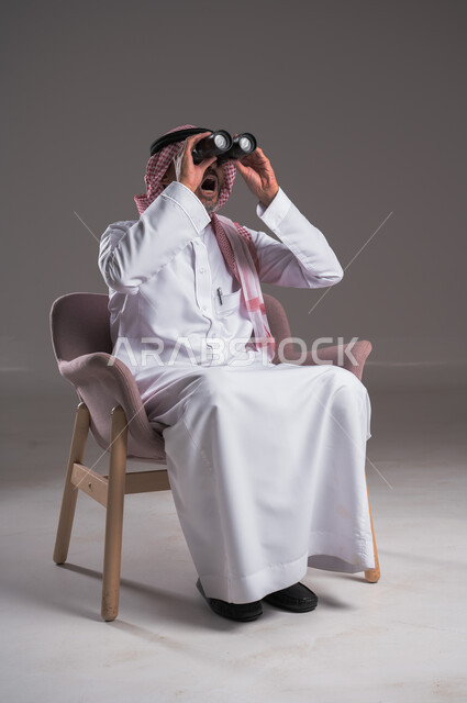 Seeing distant objects through a telescope with gestures of amazement and pleasure, planning for future goals and ambitions, portrait of a Saudi Arabian Gulf man wearing traditional thobe and shemagh sitting on a comfortable chair looking through binoculars, love of exploration and adventure, gray background