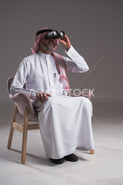الشغف وحب الاستكشاف والمغامرة، البحث عن شيء ما باستخدام الناظور، مفهوم رؤية الأشياء البعيدة، بورتريه لرجل عربي خليجي سعودي يرتدي الثوب التقليدي والشماغ يجلس على كرسي مريح  ينظر من خلال التلسكوب, حلم تحقيق الأهداف والطموحات المستقبلية, خلفية رمادية