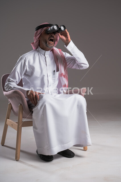 حب الاستكشاف والمغامرة، رؤية الاشياء البعيدة عبر التلسكوب بايماءات الاندهاش والسرور، التخطيط المستقبلي للأهداف والطموحات، بورتريه لرجل عربي خليجي سعودي يرتدي الثوب التقليدي والشماغ يجلس على كرسي مريح  ينظر من خلال منظار الرؤية ثنائي العينين, خلفية رمادية