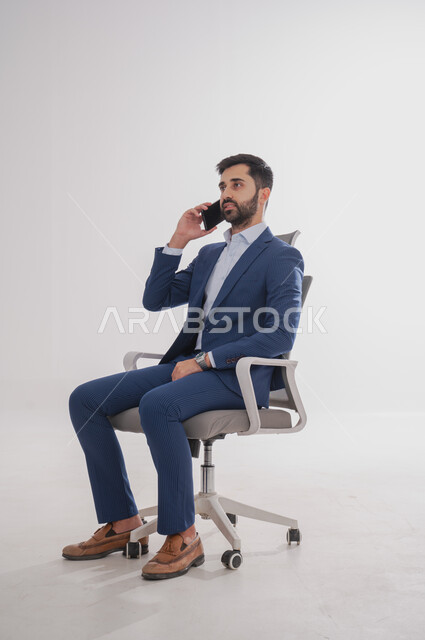 إستخدام تطبيقات الهاتف, بورتريه لرجل عربي سعودي خليجي يرتدي بدلة رسمية يجلس على كرسي  ويحمل بيده هاتف محمول بإيماءات تدل على السعادة، إجراء مكالمة صوتية, قضاء وقت ممتع، خلفية بيضاء