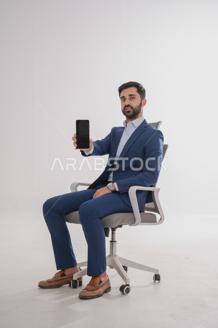 إستخدام تطبيقات الهاتف, بورتريه لرجل عربي سعودي خليجي يرتدي بدلة رسمية يجلس على كرسي  ويحمل بيده هاتف محمول بإيماءات تدل على السعادة، إجراء مكالمة صوتية, قضاء وقت ممتع، خلفية بيضاء
