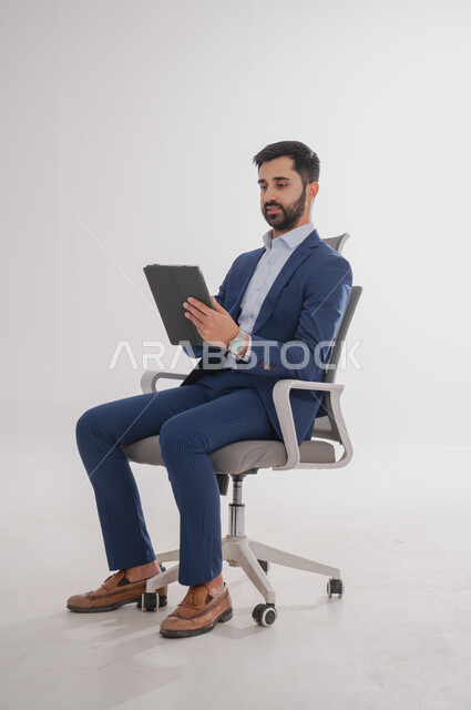 إستخدام تطبيقات الجهاز المحمول, بورتريه لرجل عربي سعودي خليجي يرتدي بدلة رسمية يجلس على كرسي  ويحمل بيده تابليت، إجراء مكالمة فيديو وصوت , قضاء وقت ممتع، خلفية بيضاء