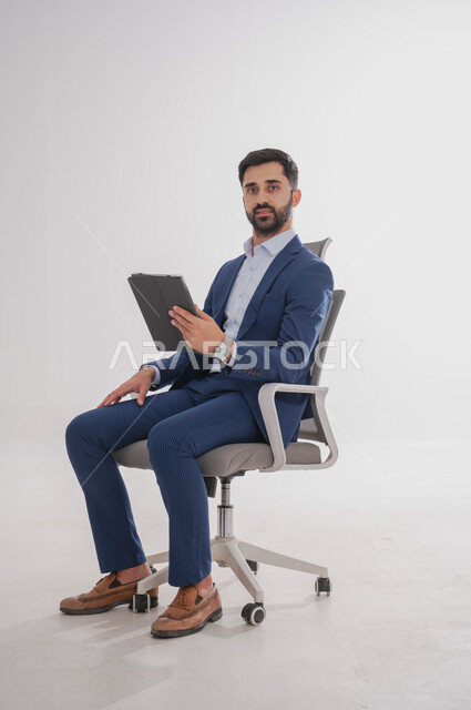إستخدام تطبيقات الجهاز المحمول, بورتريه لرجل عربي سعودي خليجي يرتدي بدلة رسمية يجلس على كرسي  ويحمل بيده تابليت، إجراء مكالمة فيديو وصوت , قضاء وقت ممتع، خلفية بيضاء