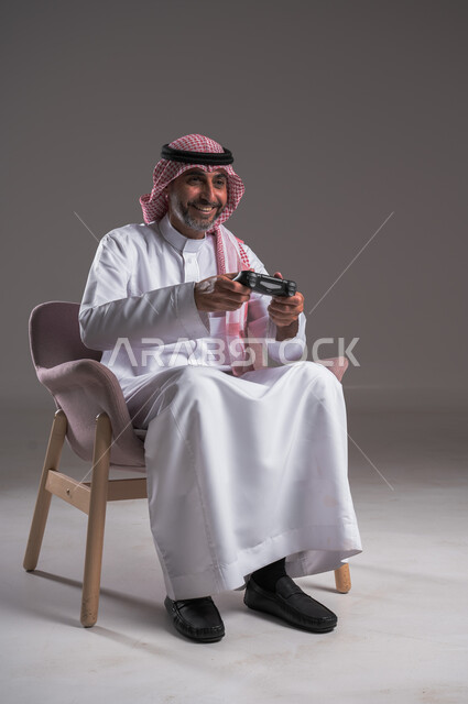 تجربة ترفيهية ممتعة ومثيرة في الأجهزة الحديثة، ايماءات الانتصار والفوز، التفاعل والاندماج مع ألعاب البلايستيشن، تقنيات الكترونية ترفيهيه, بورتريه لرجل عربي سعودي خليجي يرتدي الثوب التقليدي والشماغ يجلس على كرسي مريح يحمل بيده عصا التحكم، خلفية رمادية