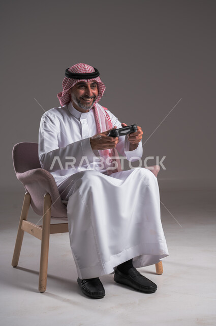 تقنيات الكترونية ترفيهيه, بورتريه لرجل عربي سعودي خليجي يرتدي الثوب التقليدي والشماغ يجلس على كرسي مريح يحمل بيده عصا التحكم، تجربة ترفيهية ممتعة ومثيرة في الأجهزة الحديثة، ايماءات الانتصار والفوز، التفاعل والاندماج مع ألعاب البلايستيشن، خلفية رمادية