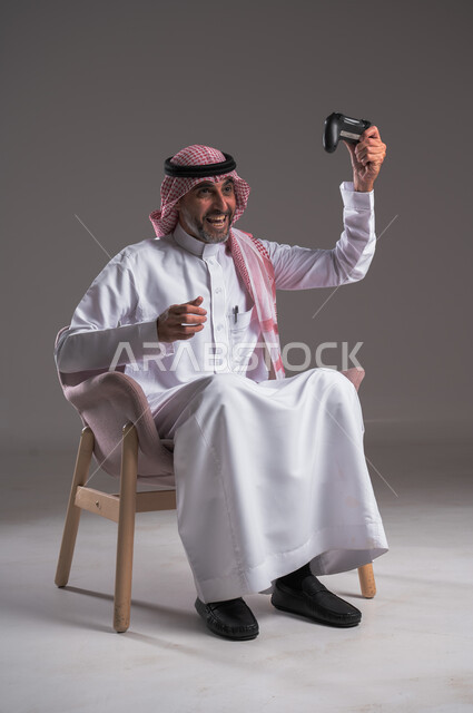 ايماءات الانتصار والفوز، التفاعل والاندماج مع ألعاب البلايستيشن، تقنيات الكترونية ترفيهيه, بورتريه لرجل عربي سعودي خليجي يرتدي الثوب التقليدي والشماغ يجلس على كرسي مريح يحمل بيده عصا التحكم، تجربة ترفيهية ممتعة ومثيرة في الأجهزة الحديثة، خلفية رمادية