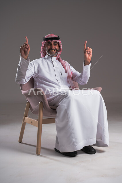 تحديد موقع معين بايماءات السعادة والسرور، إشارات وحركات يدين تدل على هذا المكان (هنا)، الإشارة والتوضيح إلى شيء ما، بورتريه لرجل عربي خليجي سعودي يرتدي الثوب التقليدي والشماغ يجلس على كرسي مريح ينظر إلى الكاميرا ويرفع اصبعه السبابة إلى الأعلى، خلفية رمادية