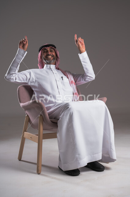 الإشارة والتوضيح إلى شيء ما، بورتريه لرجل عربي خليجي سعودي يرتدي الثوب التقليدي والشماغ يجلس على كرسي مريح ينظر إلى الكاميرا ويرفع اصبعه السبابة إلى الأعلى، تحديد موقع معين بايماءات السعادة والسرور، إشارات وحركات يدين تدل على هذا المكان (هنا)، خلفية رمادية
