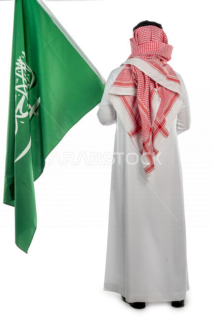 تصوير من الخلف لرجل عربي خليجي سعودي يرتدي الزي الخليجي التقليدي، بيده علم المملكة العربية السعودية ، ذكرى اليوم الوطني السعودي ،الاحتفال بالعيد الوطني السعودي