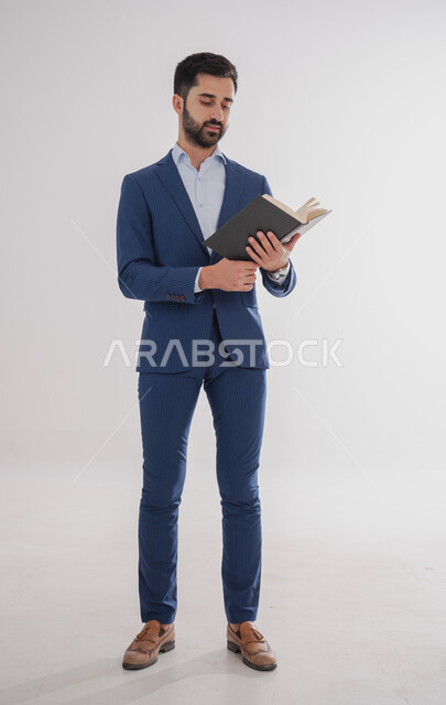 مفهوم المطالعة والتعلم, قضاء وقت كافي للتثقف, بورتريه لرجل عربي سعودي خليجي يرتدي بدلة رسمية يقف ويمسك بيده كتاب للقراءة, الجسم بالطول الكامل, خلفية بيضاء