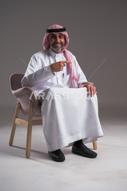 النظر الكاميرا بإيماءات السرور والبشاشة، بورتريه لرجل عربي خليجي سعودي مبتسم يرتدي الزي التقليدي والشماغ يجلس على كرسي مريح و يشير بأصابعه الى هدف محدد، إشارات وحركات يدين تدل على هذا المكان (هنا)، تحديد موقع معين، خلفية رمادي