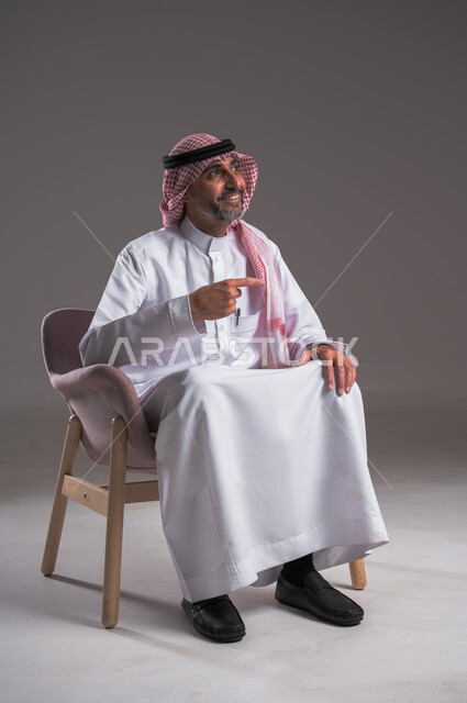 إشارات وحركات يدين تدل على هذا المكان (هنا)، تحديد موقع معين، النظر الى شيء ما بإيماءات السرور والبشاشة، بورتريه لرجل عربي خليجي سعودي مبتسم يرتدي الزي التقليدي والشماغ يجلس على كرسي مريح و يشير بأصابعه الى هدف محدد، خلفية رمادية