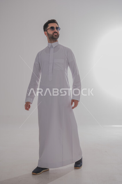 مفهوم الاناقة والشياكة, الاهتمام بالمظهر الخارجي, بورتريه لرجل عربي سعودي خليجي يرتدي الثوب ويقف بايماءات الثقة بالنفس, الجسم بالطول الكامل, خلفية بيضاء
