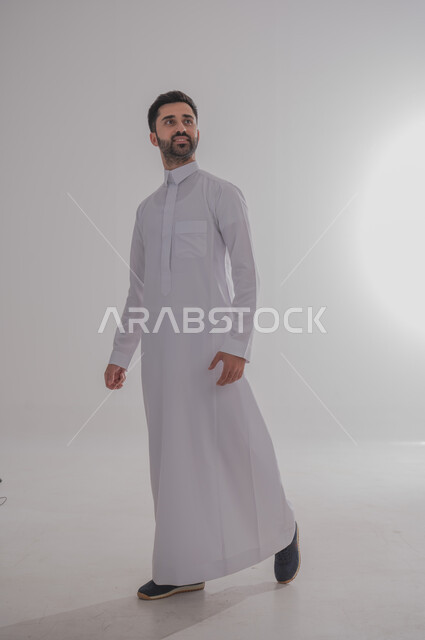 مفهوم الاناقة والشياكة, الاهتمام بالمظهر الخارجي, بورتريه لرجل عربي سعودي خليجي يرتدي الثوب ويقف بايماءات الثقة بالنفس, الجسم بالطول الكامل, خلفية بيضاء