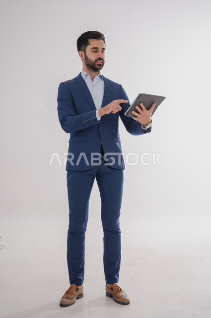 Using modern technical devices, a portrait of a Saudi Gulf Arab man wearing a formal suit and holding a tablet in his hand, chatting with family and friends, browsing social media programs, tablet applications, full-length body photo, white background.