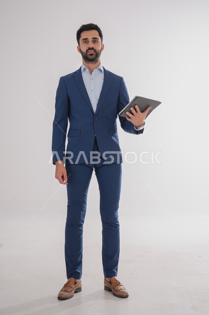 استخدام الأجهزة التقنية الحديثة, بورتريه لرجل عربي خليجي سعودي يرتدي  البدلة الرسمية ويحمل بيده التابليت، الدردشة مع الأهل والأصدقاء, تصفح برامج التواصل الاجتماعي، تطبيقات الأجهزة اللوحية، صورة الجسم بالطول الكامل, خلفية بيضاء