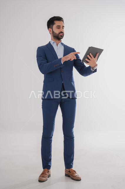 استخدام الأجهزة التقنية الحديثة, بورتريه لرجل عربي خليجي سعودي يرتدي  البدلة الرسمية ويحمل بيده التابليت، الدردشة مع الأهل والأصدقاء, تصفح برامج التواصل الاجتماعي، تطبيقات الأجهزة اللوحية، صورة الجسم بالطول الكامل, خلفية بيضاء