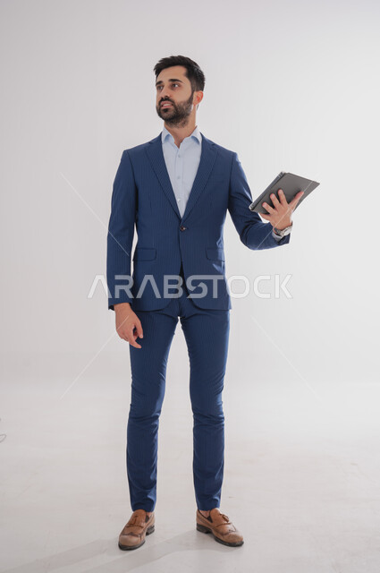 استخدام الأجهزة التقنية الحديثة, بورتريه لرجل عربي خليجي سعودي يرتدي  البدلة الرسمية ويحمل بيده التابليت، الدردشة مع الأهل والأصدقاء, تصفح برامج التواصل الاجتماعي، تطبيقات الأجهزة اللوحية، صورة الجسم بالطول الكامل, خلفية بيضاء