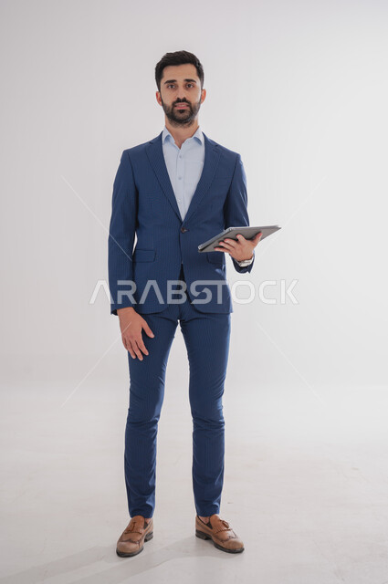 استخدام الأجهزة التقنية الحديثة, بورتريه لرجل عربي خليجي سعودي يرتدي  البدلة الرسمية ويحمل بيده التابليت، الدردشة مع الأهل والأصدقاء, تصفح برامج التواصل الاجتماعي، تطبيقات الأجهزة اللوحية، صورة الجسم بالطول الكامل, خلفية بيضاء