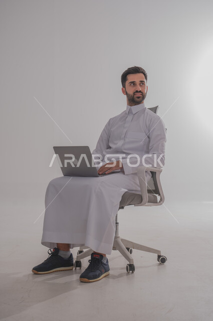Using modern technical devices, a portrait of a Saudi Gulf Arab man wearing a thobe, sitting on a chair and placing a laptop in front of him, chatting with family and friends, browsing social media programs, computer applications, white background.