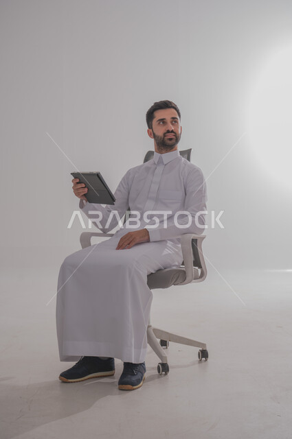 استخدام الأجهزة التقنية الحديثة, بورتريه لرجل عربي خليجي سعودي يرتدي  الثوب ويجلس على كرسي ويحمل بيده تابليت، الدردشة مع الأهل والأصدقاء, تصفح برامج التواصل الاجتماعي، تطبيقات الأجهزة اللوحية،  خلفية بيضاء