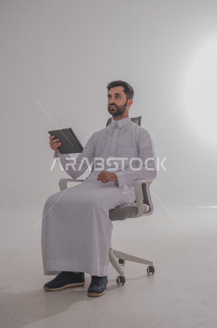 استخدام الأجهزة التقنية الحديثة, بورتريه لرجل عربي خليجي سعودي يرتدي  الثوب ويجلس على كرسي ويحمل بيده تابليت، الدردشة مع الأهل والأصدقاء, تصفح برامج التواصل الاجتماعي، تطبيقات الأجهزة اللوحية،  خلفية بيضاء