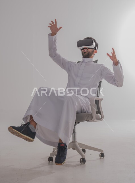 إيماءات الاندماج والتفاعل مع العالم الخيالي، بورتريه  لرجل عربي خليجي سعودي يرتدي الثوب يجلس ويضع نظارة الفي آر ثلاثية الأبعاد، محاكاة الواقع الافتراضي باستخدام التقنيات الحديثة المتطورة، خلفية بيضاء
