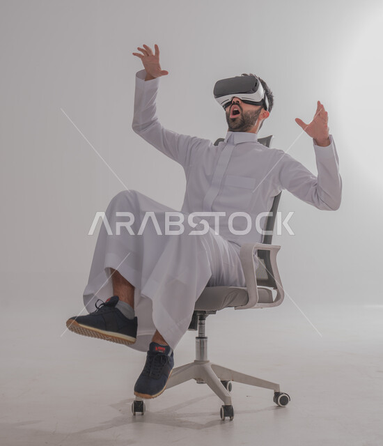 إيماءات الاندماج والتفاعل مع العالم الخيالي، بورتريه  لرجل عربي خليجي سعودي يرتدي الثوب يجلس ويضع نظارة الفي آر ثلاثية الأبعاد، محاكاة الواقع الافتراضي باستخدام التقنيات الحديثة المتطورة، خلفية بيضاء