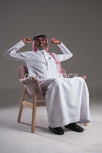 ايماءات وجه ويدين تدل على الراحة، النظر الى شيء ما بتعابير التفكيروشرود الذهن، الشعور بالتعب والحاجة إلى النوم، مفهوم الاسترخاء والاستمتاع بالأجواء الهادئة، بورتريه لرجل عربي خليجي سعودي يرتدي الثوب التقليدي والشماغ يجلس على كرسي مريح، خلفية رمادية 