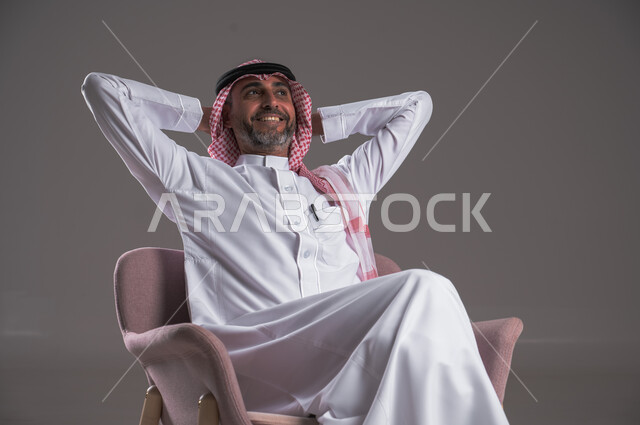 الشعور بالتعب والحاجة إلى النوم، مفهوم الاسترخاء والاستمتاع بالأجواء الهادئة، بورتريه لرجل عربي خليجي سعودي يرتدي الثوب التقليدي والشماغ يجلس على كرسي مريح ويضع يديه تحت رأسه، تعابير وإيماءات الراحة والشرود، النظر الى شيء ما، خلفية رمادية 