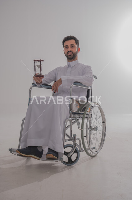 ذوي الاحتياجات الخاصة والهمم، بورتريه  لرجل عربي خليجي سعودي يرتدي الثوب يجلس على كرسي متحرك ويحمل بيده ساعة رملية، خلفية بيضاء