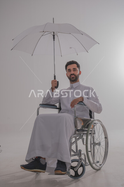 Winter and rainy weather, people with special needs and concerns, portrait of a Saudi Gulf Arab man wearing a thobe, sitting in a wheelchair and holding an umbrella in his hand, white background
