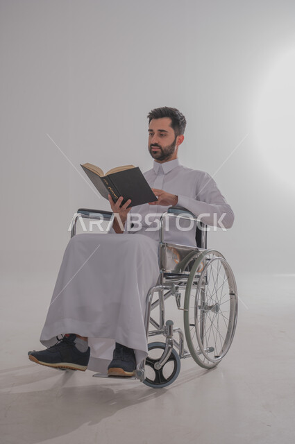 مفهوم المطالعة والتثقف والقراءة, ذوي الاحتياجات الخاصة والهمم، بورتريه  لرجل عربي خليجي سعودي يرتدي الثوب يجلس على كرسي متحرك ويحمل بيده كتاب، خلفية بيضاء