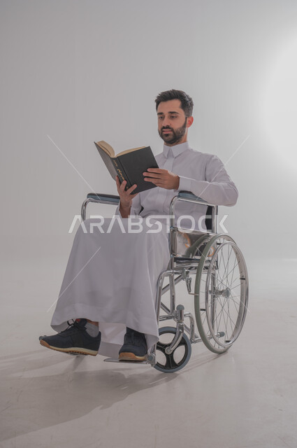 مفهوم المطالعة والتثقف والقراءة, ذوي الاحتياجات الخاصة والهمم، بورتريه  لرجل عربي خليجي سعودي يرتدي الثوب يجلس على كرسي متحرك ويحمل بيده كتاب، خلفية بيضاء