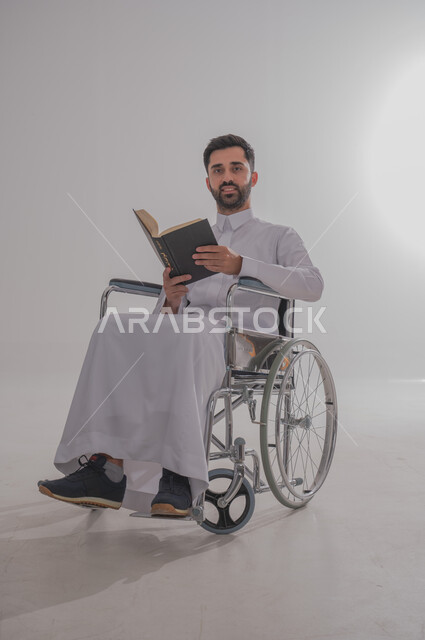 مفهوم المطالعة والتثقف والقراءة, ذوي الاحتياجات الخاصة والهمم، بورتريه  لرجل عربي خليجي سعودي يرتدي الثوب يجلس على كرسي متحرك ويحمل بيده كتاب، خلفية بيضاء