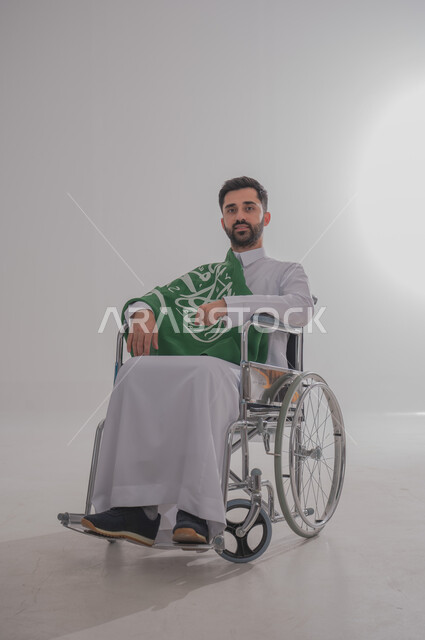 مفهوم الاحتفال باليوم الوطني, ذوي الاحتياجات الخاصة والهمم، بورتريه  لرجل عربي خليجي سعودي يرتدي الثوب يجلس على كرسي متحرك ويضع على كتفه علم المملكة العربية السعودية، حب الوطن والفخر والاعتزاز به,  خلفية بيضاء
