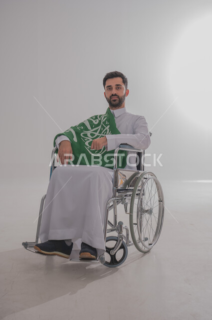 مفهوم الاحتفال باليوم الوطني, ذوي الاحتياجات الخاصة والهمم، بورتريه  لرجل عربي خليجي سعودي يرتدي الثوب يجلس على كرسي متحرك ويضع على كتفه علم المملكة العربية السعودية، حب الوطن والفخر والاعتزاز به,  خلفية بيضاء