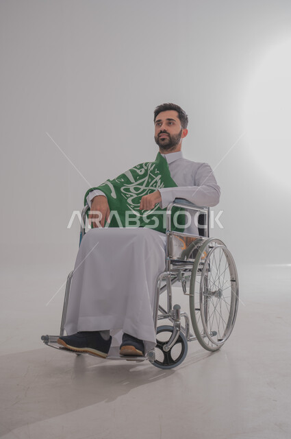 مفهوم الاحتفال باليوم الوطني, ذوي الاحتياجات الخاصة والهمم، بورتريه  لرجل عربي خليجي سعودي يرتدي الثوب يجلس على كرسي متحرك ويضع على كتفه علم المملكة العربية السعودية، حب الوطن والفخر والاعتزاز به,  خلفية بيضاء
