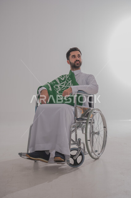 مفهوم الاحتفال باليوم الوطني, ذوي الاحتياجات الخاصة والهمم، بورتريه  لرجل عربي خليجي سعودي يرتدي الثوب يجلس على كرسي متحرك ويضع على كتفه علم المملكة العربية السعودية، حب الوطن والفخر والاعتزاز به,  خلفية بيضاء