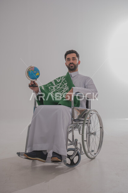مفهوم الاحتفال باليوم الوطني, ذوي الاحتياجات الخاصة والهمم، بورتريه  لرجل عربي خليجي سعودي يرتدي الثوب يجلس على كرسي متحرك يضع على كتفه علم المملكة العربية السعودية ويحمل بيده كرة ارضية، حب الوطن والفخر والاعتزاز به,  خلفية بيضاء