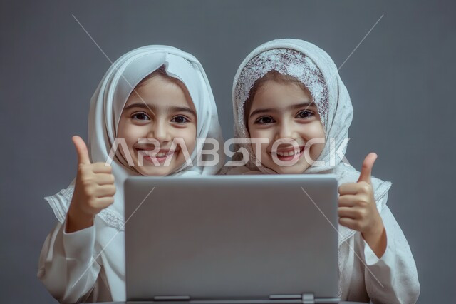 متابعة حصص الاونلاين عن طريق جهاز الحاسوب، بورتريه لطالبتان عربيتان خليجيتان سعوديتان محجبتان يستخدمان الحاسب المحمول وينظران إلى الكاميرا بإيماءات السعادة والسرور يرفعن اصبع الابهام الى الاعلى، التعلم عن بعد باستخدام التكنولوجيا الحديثة، خلفية بيضاء