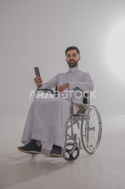 النظر إلى الكاميرا بإيماءات البشاشة، الاستفادة التقنيات الحديثة والتكنولوجيا المتطورة، بورتريه لشاب عربي خليجي سعودي من ذوي الاحتياجات الخاصة يرتدي الثوب التقليدي يجلس على كرسي متحرك يمسك الهاتف المحمول بيده، تصفح مواقع الانترنت ومتابعة الأخبار، خلفية رمادية