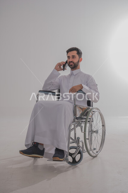إجراء مكالمات هاتفية باستخدام جهاز الجوال، الاستفادة التقنيات الحديثة والتكنولوجيا المتطورة، بورتريه لشاب عربي خليجي سعودي من ذوي الاحتياجات الخاصة يرتدي الثوب التقليدي يجلس على كرسي متحرك يستخدم الهاتف المحمول، خلفية رمادية