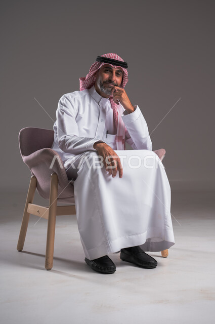 الاهتمام بالأناقة والمظهر الخارجي، بورتريه لرجل عربي خليجي سعودي يرتدي الثوب التقليدي والشماغ يجلس على كرسي مريح ويضع يده تحت ذقنه، النظر إلى الكاميرا بإيماءات التركيز، التمعن والتخطيط للمستقبل, خلفية رمادية 