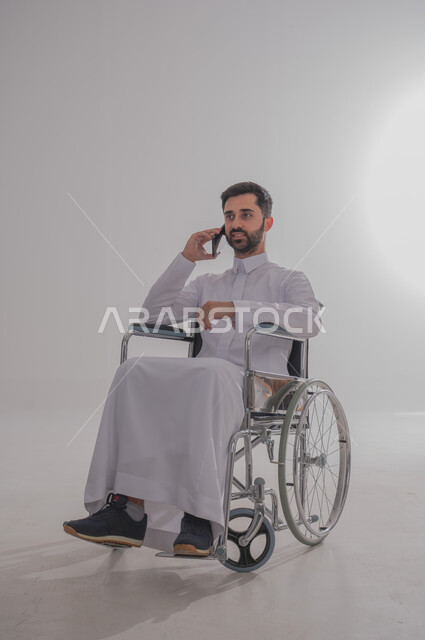 إجراء المكالمات الهاتفية باستخدام جهاز الجوال، الاستفادة التقنيات الحديثة والتكنولوجيا المتطورة، بورتريه لشاب عربي خليجي سعودي من ذوي الاحتياجات الخاصة يرتدي الثوب التقليدي يجلس على كرسي متحرك يستخدم الهاتف المحمول، خلفية رمادية