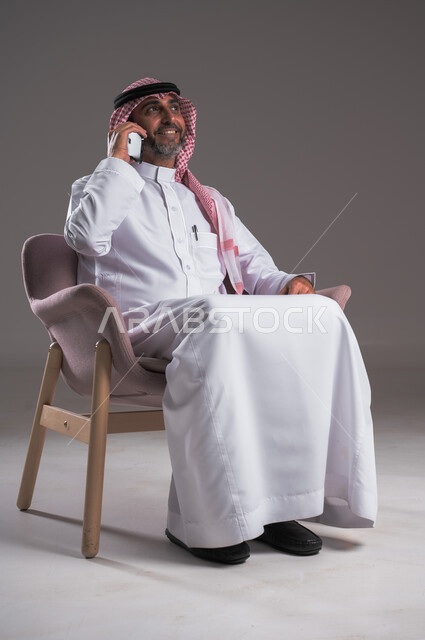 استخدام الوسائل التقنية الحديثة في الحياة العملية، الشعور بالسعادة لسماع خبر ما، إجراء مكالمات هاتفية عبر الهاتف المحمول, التواصل مع الأهل والأصدقاء، بورتريه لرجل عربي سعودي خليجي مبتسم يرتدي الثوب التقليدي والشماغ يجلس على كرسي مريح ويستخدم الجوال، خلفية رمادية